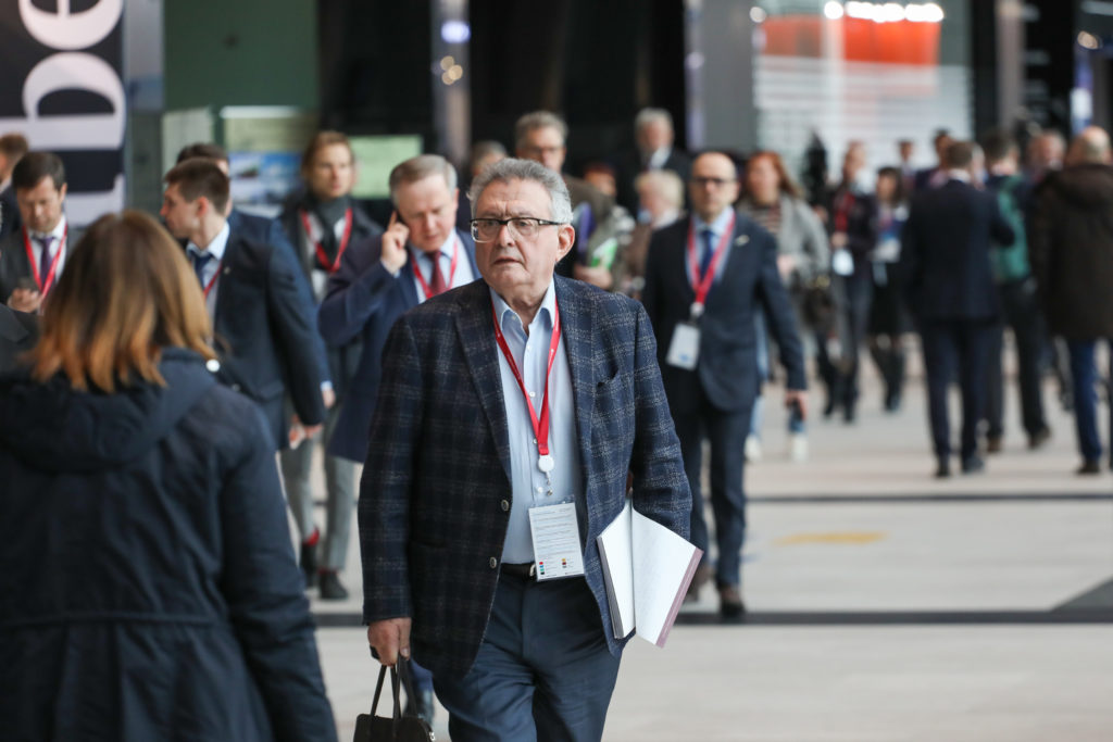 Participants with badges walk through a forum corridor, with a man in a suit in the center.