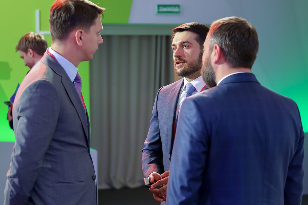 Several men in suits talk in a hall with green lighting.