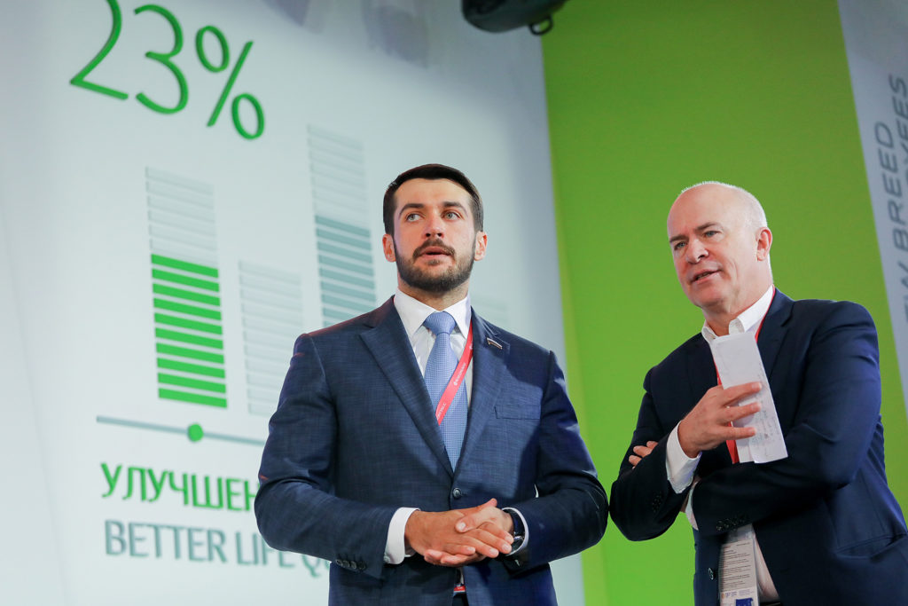 Two men in suits stand on a stage in front of a green screen with a chart.